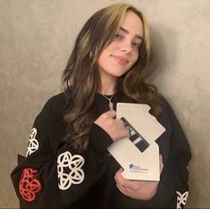 a woman is holding an item in her right hand and smiling at the camera while standing against a wall