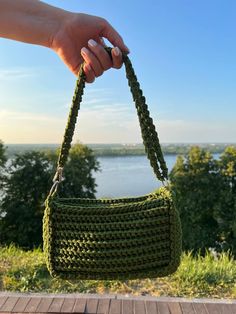 a hand holding a green crocheted purse over a wooden deck overlooking a body of water