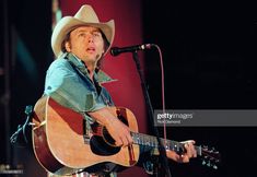 a man wearing a cowboy hat and holding a guitar in front of a microphone on stage