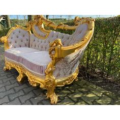 a gold and white couch sitting on top of a brick floor next to bushes in front of a hedge