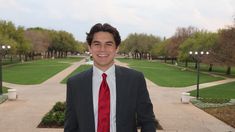 a man wearing a suit and tie standing in front of a park