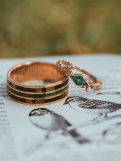 A custom-made 14K rose gold engagement ring from Staghead Designs sits on a book page. This ring, called "Sparrow," has an east-west set kite cut lab-created emerald center stone, leaf accent metalwork, & a metal sparrow embellishment. This ring can also be made in 14K yellow or white gold & with a variety of unique gemstones. Find or design your dream wedding rings today with Staghead Designs! Emerald Stone Engagement Ring, Sparrow Ring, Lesbian Engagement Ring, Lozenge Cut, Antler Wedding Rings, Rose Quartz Ring Engagement, Engagement Rings Wedding Bands Set, Dream Wedding Ring, Wood Wedding Ring