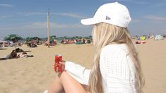 a woman sitting on top of a sandy beach next to a red can filled with liquid