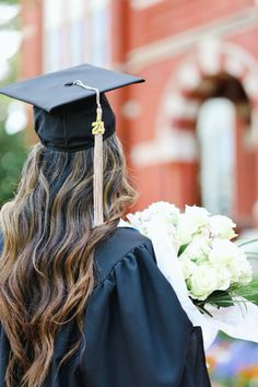 a woman wearing a graduation cap and gown holding flowers in front of a brick building