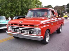 an old red truck is driving down the street with other classic cars in the background