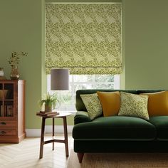 a green couch sitting under a window next to a table with a lamp on it