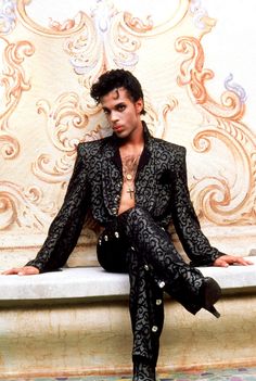 a man sitting on top of a white bench wearing black pants and a suit jacket