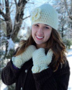 a woman in winter clothing holding onto her mittens and smiling at the camera with snow covered trees behind her