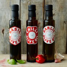 three bottles of garlic oil next to some red peppers and an onion on a wooden table