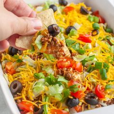 a hand dipping a tortilla chip into a casserole dish with black olives, lettuce and tomatoes