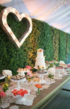 a table topped with lots of cake and cupcakes next to a wall covered in greenery