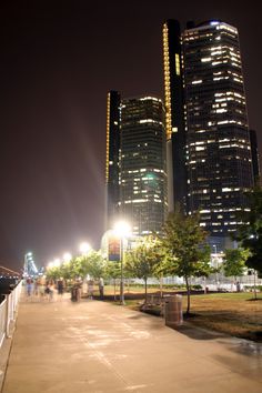 people are walking on the sidewalk in front of tall buildings at night with bright lights