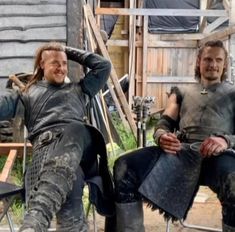 two men sitting on top of chairs in front of a wooden building and one man with long hair