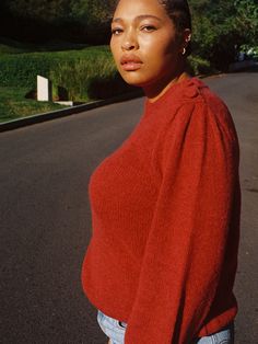 a woman standing on the side of a road wearing a red sweater and blue jeans