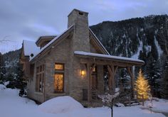 a small stone house with a light on in the front yard covered in snow and surrounded by evergreen trees