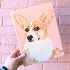 a hand holding up a painting of a dog on a pink brick wall with the face of a corgi