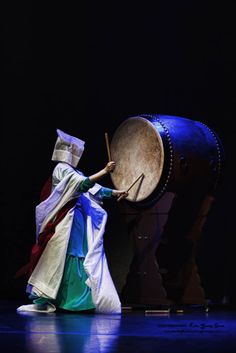 a person in costume is playing with a large drum on a dark stage, while wearing a white hat and cloak