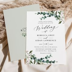 two wedding cards with greenery are laying on a fur - lined bed sheet, next to each other