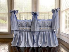 a baby crib with blue ruffles and bows on the top, in front of windows