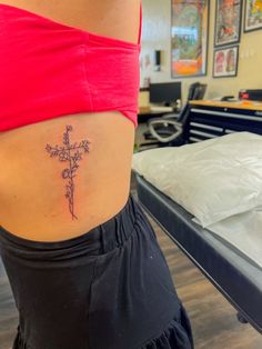 a woman with a cross tattoo on her lower back side ribcage, standing in front of a bed