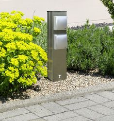 there is a metal box on the side of the road next to some plants and flowers