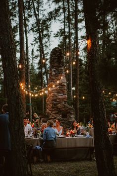 a group of people sitting around a table in the woods with lights strung over them