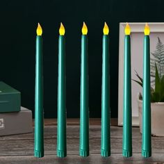 five green candles are lined up next to each other on a table with a plant in the background