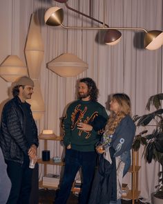 three people standing in a living room talking to each other and two lamps hanging from the ceiling