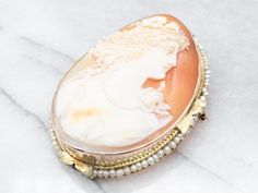 "This cameo features wonderful, crisp details, and a great contrast between the creamy white carving and the soft russet of the background. A string of seed pearls pulls the piece together, drawing on the color of the profile and the floral details of the gold. Metal: 14K Yellow Gold Gem: Shell Cameo Gem Measurements: 34.4 x 45.6 mm, Oval Accents: Seed Pearls Measurements: 40 x 55 mm Marks: \"X\" Stamped To view a video of this piece check out the link below: https://vimeo.com/802860432 SKU #: A Antique White Brooch For Formal Occasions, Exquisite White Brooch For Formal Occasions, Antique White Formal Brooch, Luxury Intaglio Brooch For Formal Occasions, Elegant Formal Brooch With Intaglio Detail, Elegant Intaglio Brooch For Formal Occasions, Elegant Cream Brooches For Formal Occasions, White Cameo Brooch For Formal Occasions, White Cameo Brooches For Formal Occasion