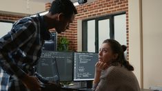 a man and woman looking at two computer monitors in an office setting with brick walls