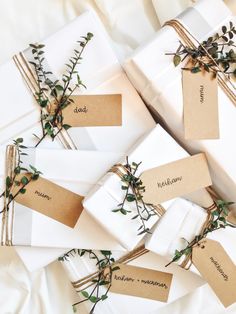 wrapped presents with tags tied to them on a white cloth covered tablecloth, surrounded by greenery and twine