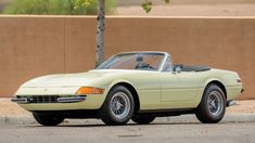 an old yellow sports car parked in front of a tan wall with trees on it