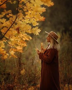 a woman wearing a hat standing in front of a tree with yellow leaves on it