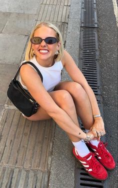 a woman sitting on the ground with her legs crossed and wearing red tennis shoes, holding a black purse