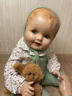 a baby doll sitting on the floor with a teddy bear in front of it's face