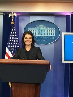 a woman standing at a podium in front of a white house sign and american flag