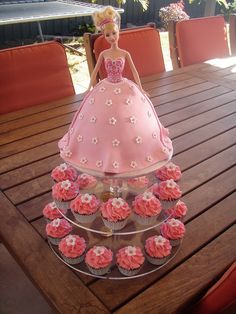 a barbie doll cake and cupcakes on a wooden table with chairs in the background