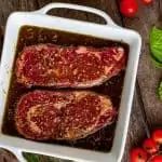 two steaks in a white casserole dish with tomatoes and basil on the side