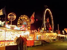 people standing in front of carnival booths at night with ferris wheel and flags on the sides