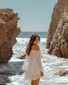 a beautiful woman standing on top of a beach next to the ocean wearing a white dress