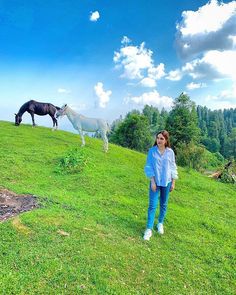 a woman standing on top of a lush green hillside next to two horses in the distance