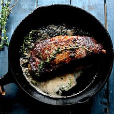 a piece of meat in a skillet on a blue wooden table with some herbs