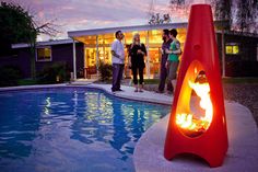 three people standing around a fire pit next to a swimming pool at night with the lights on