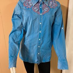 New Grace Buttoned Down Women's Blouse. Material Is Turquoise Blue With A Worn-In Look. Fabric Is Regular Weight. A Little Bling Within The Pink Embroidered Flowers. Tailored To Show Off Your Figure. Hem Length (From Collar) = 29". Wear Tucked In Or Out. Back Also Has Designed Yoke. Comes In Medium, Large And Extra Large. Denim Blue Long Sleeve Tops With Floral Embroidery, Western Style Blue Tops For Spring, Western Blue Button-up Top, Blue Western Tops For Fall, Blue Western Style Tops For Fall, Fitted Embroidered Medium Wash Tops, Embroidered Long Sleeve Denim Shirt, Embroidered Blue Denim Blouse, Blue Denim Tops With Floral Embroidery