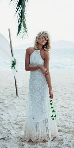 a woman is standing on the beach in a white wedding dress and holding a bouquet