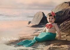 a baby mermaid sits on the beach with her tail up and looks at the camera