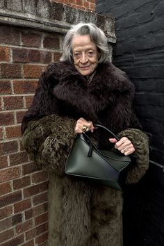 an older woman in a fur coat holding a green handbag while leaning against a brick wall