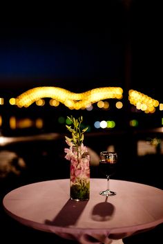 a vase with flowers on top of a table next to a wine glass and some lights in the background