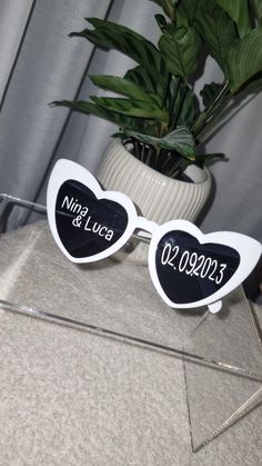 two heart shaped sunglasses with the names and date on them, sitting next to a potted plant