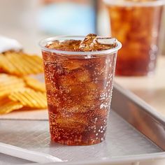 a plastic cup filled with soda next to some crackers and chips on a tray